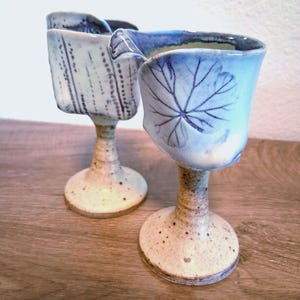 Puede incluir: Dos copas de cerámica hechas a mano. Una copa presenta un diseño de hoja, la otra tiene un patrón de puntos. Ambas tienen una base y un tallo beige moteado, con detalles en azul y blanco. Las copas están sobre una superficie de madera.