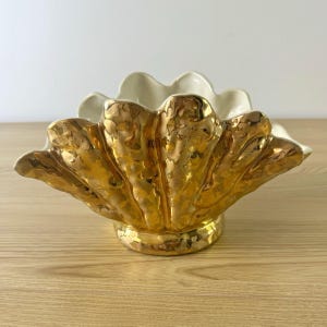 May include: A ceramic bowl shaped like a large seashell with a metallic gold exterior finish and a smooth cream-colored interior. The bowl features a scalloped edge and a textured, rippled surface that mimics the natural ridges of a shell.