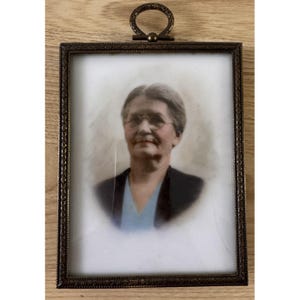May include: A rectangular portrait frame with an ornate, dark metallic finish and a circular hanging loop at the top. The frame contains a vintage-style photograph of a person with short hair and round glasses, wearing a dark jacket over a blue shirt.