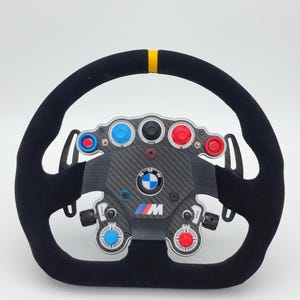 Op de afbeelding: Een zwart racestuur met een gele streep aan de bovenkant. Het heeft een koolstofvezel middenstuk met gekleurde knoppen en het BMW-logo. Het stuur heeft een suède-achtige textuur.