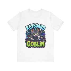 May include: White t-shirt featuring a cartoon cat typing on a keyboard. The cat is gray and white with a grumpy expression. The text "KEYBOARD GOBLIN" is above and below the cat in blue and green.