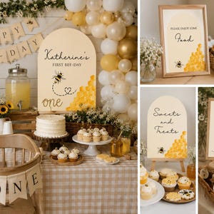 May include: A bee-themed first birthday party setup with decorations, including a banner, signs, and a cake. The decorations feature bee and honeycomb motifs, with a color scheme of yellow, white, and beige.