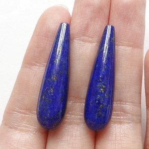 Può includere: Due pietre preziose allungate di lapislazzuli a forma di goccia. Le pietre blu scuro hanno macchie d'oro e sono tenute in una mano su sfondo bianco. Queste pietre lucide sono probabilmente per la creazione di gioielli.
