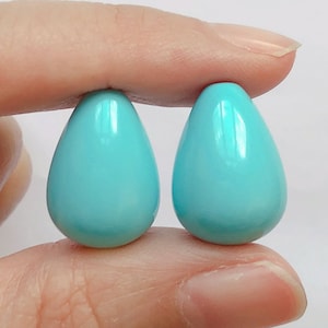 May include: Two teardrop-shaped turquoise blue beads, held between two fingers.