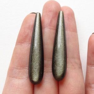 Può includere: Due pendenti allungati a forma di goccia, dall'aspetto marrone dorato scintillante. I pendenti sono tenuti in mano su sfondo bianco, mostrando la loro superficie lucida e il loro colore unico. Questi pendenti sono probabilmente per la creazione di gioielli.