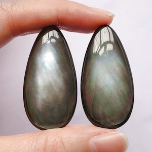May include: Two iridescent, teardrop-shaped gemstones. The stones have a dark, smoky base color with shimmering, pearlescent highlights. The smooth, polished surfaces reflect light, creating a play of colors. The gemstones are held against a light background.