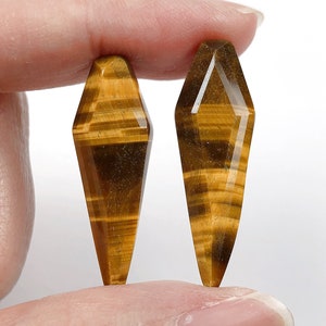 May include: Two golden brown tiger's eye gemstone points, each with a unique pattern of stripes, are held between two fingers.