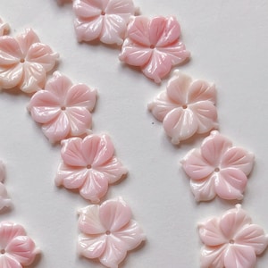 Può includere: Una collezione di perline a forma di fiore di conchiglia rosa e bianca. Le perline sono disposte in un motivo circolare su uno sfondo bianco.