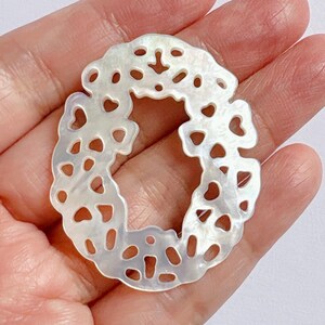 May include: A pearlescent, open-worked pendant in the shape of the letter "O". The pendant has a delicate, lacy design with heart-shaped and circular cutouts. The iridescent surface reflects light, creating a shimmering effect.