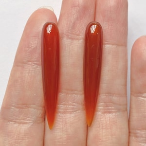 May include: Two long, pointed, red carnelian stones. The stones are smooth and polished, with a slight sheen.