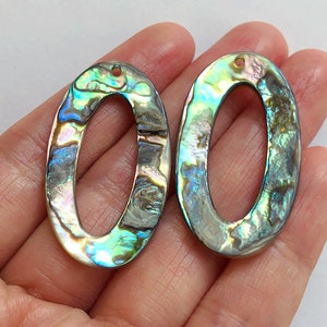 Può includere: Due ciondoli ovali in madreperla di abalone con un foro nella parte superiore per l'appeso. Le conchiglie hanno un colore blu, verde e marrone iridescente.