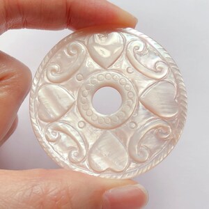 May include: A white mother of pearl pendant with a carved design of hearts and swirls. The pendant has a hole in the center for a cord or chain.