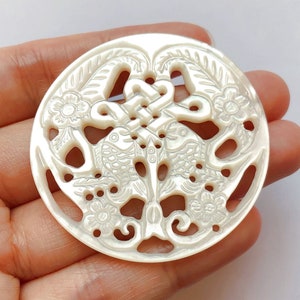 Może przedstawiać: Okrągły wisiorek z białej perłowej muszli z rzeźbionym wzorem dwóch ryb i węzła. Wisiorek jest okrągły i ma delikatny, skomplikowany wzór.