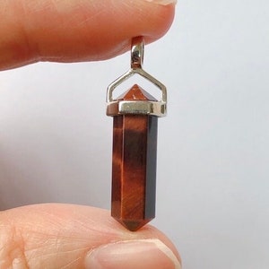 May include: A red tiger's eye crystal point pendant with a silver setting. The pendant is about 2 inches long and is held by a finger.