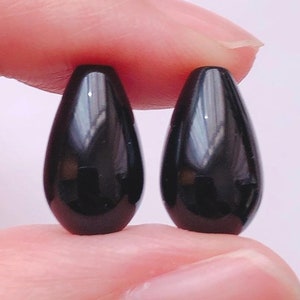 Può includere: Due perline nere a forma di goccia, tenute tra due dita.