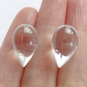 May include: Two clear, teardrop-shaped glass beads. Each bead has a pointed bottom and a rounded top. The beads are held in a person's hand against a white background. The beads are transparent and reflect light.