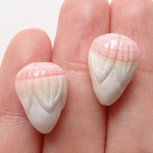 Peut inclure: Deux petites perles en coquillage sculpté en forme de goutte, dotées d'une finition lisse et polie. Chaque perle présente un dégradé de rose tendre sur le sommet arrondi, passant au blanc crème à la base pointue avec des détails sculptés.