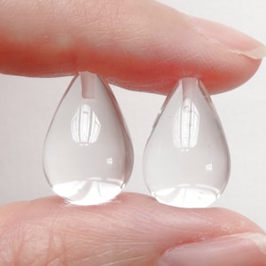 Può includere: Due perline di vetro trasparente a forma di lacrima, tenute tra due dita.