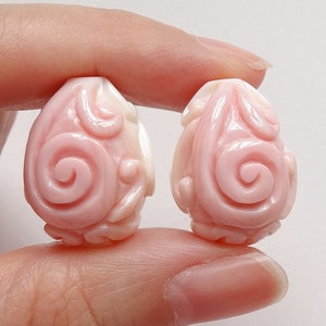 May include: Two pink and white shell beads with a carved floral design. The beads are oval shaped and have a swirl pattern in the center.