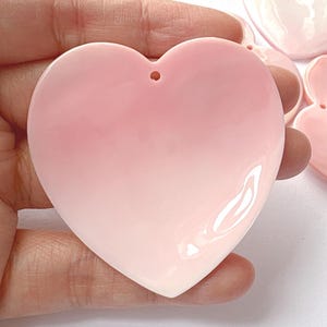 May include: A large, glossy, heart-shaped pendant in shades of pink, held in a hand. The heart has a hole at the top for stringing. Other similar pendants are visible in the background. The pendant is likely for jewellery making or crafting.
