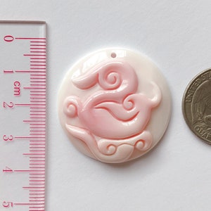 May include: A pink and white shell pendant with a carved design of a stylized bird. The pendant is approximately 2.5 cm in diameter.
