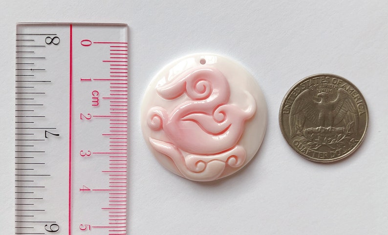 May include: A pink and white shell pendant with a carved design of a stylized bird. The pendant is approximately 2.5 cm in diameter.