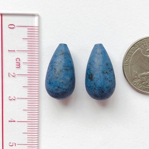 May include: Two teardrop-shaped blue beads with a speckled texture, next to a US quarter for size comparison.