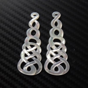 Może przedstawiać: Dwa białe wisiorki z perłowej muszli z celtyckim wzorem węzła. Wisiorki są misterne rzeźbione i mają perłowy połysk.