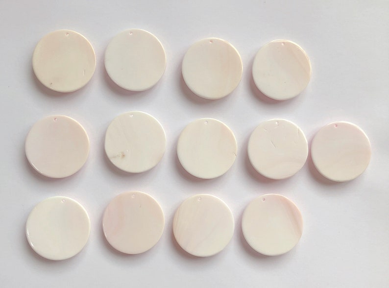 May include: Twelve white round shell pendants with a hole in the center. The pendants are arranged in three rows of four.