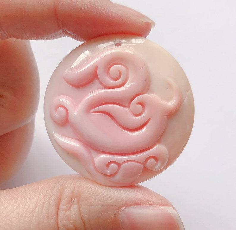 May include: A pink shell pendant with a carved design of a bird with swirling patterns. The pendant is round and has a hole at the top for hanging.