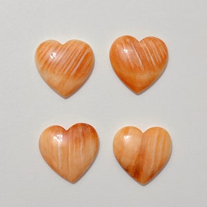 May include: Four heart-shaped cabochons made of polished, light brown, and white banded shell. The cabochons have a smooth, glossy finish.