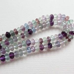 Könnte beinhalten: Eine Strähne facettierter Fluoritperlen in Violett-, Grün- und Blautönen. Die Perlen sind auf einem durchsichtigen Faden aufgefädelt und haben einen Durchmesser von etwa 6 mm.