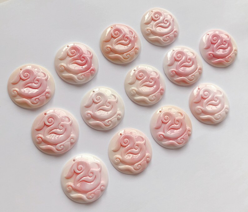 May include: A collection of 12 pink and white shell pendants with intricate carvings of clouds and a bird. The pendants are arranged in a circle on a white background.