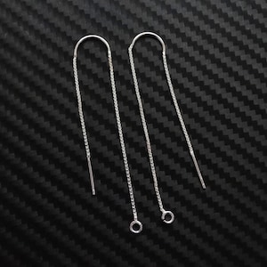 May include: A pair of silver threader earrings. Each earring features a delicate chain that hangs from a curved top, with a small bar at the end. The earrings are displayed against a textured black background.