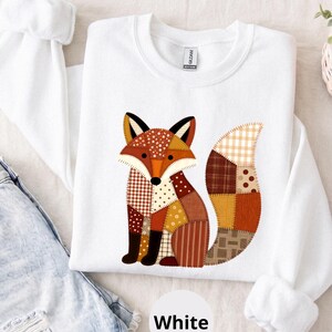 May include: A white long-sleeve sweatshirt featuring a central graphic of a fox composed of various brown, orange, and cream-colored fabric patterns including plaid, polka dots, and stripes. The fox is depicted in a seated position with a large tail.