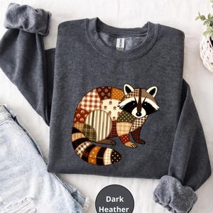 May include: A dark heather gray crewneck sweatshirt featuring a graphic of a raccoon composed of various brown, tan, and cream-colored patchwork patterns including plaid, stripes, and floral prints. The sweatshirt has long sleeves with ribbed cuffs.