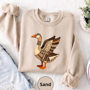 May include: A sand-coloured crewneck sweatshirt featuring a graphic of a goose designed with a patchwork pattern in various shades of brown, tan, and cream. The goose has bright orange feet and a beak, with a Gildan brand tag visible at the neck.