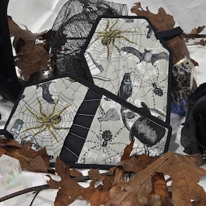 May include: A coffin-shaped fabric pouch featuring a light gray background with a black and gold spider web, spider, and bat print. The pouch has black fabric trim and is surrounded by dried brown leaves, a black rose, and various crystals.
