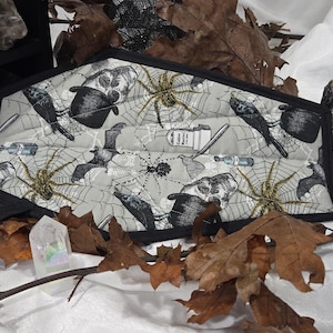 May include: A pentagon-shaped fabric pot holder featuring a light gray background with a repeating pattern of black spiders, spiderwebs, bats, skulls, top hats, and tombstones, all finished with a solid black fabric border and a small hanging loop.