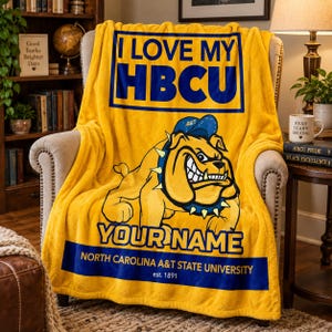 Könnte beinhalten: Eine leuchtend gelbe Fleecedecke mit dem blauen Schriftzug I LOVE MY HBCU, einer Grafik einer Bulldogge mit blauer Mütze und Stachelhalsband, dem Text YOUR NAME sowie North Carolina A&T State University est. 1891 in blauem Text.