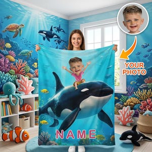 Puede incluir: Una manta de forro polar personalizada que muestra una orca grande de color blanco y negro nadando en un océano azul vibrante. Una ilustración estilo caricatura de un niño aparece montada sobre el lomo de la orca. La palabra NAME está en letras rosas.