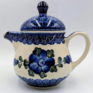 May include: A ceramic teapot with a cream-colored base featuring a hand-painted blue floral design in the center. The lid, rim, and base are decorated with a dark blue and white patterned border. It has a curved handle and a short spout.