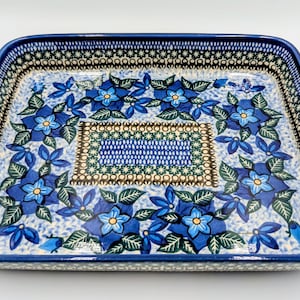 May include: A rectangular ceramic baking dish with a blue floral design. The dish features a dark blue border with a repeating pattern. The interior is decorated with blue flowers and green leaves, with a central rectangular design.
