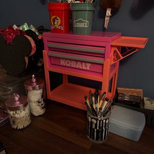 May include: A bright pink and orange Kobalt metal tool chest with two drawers and a side tray. On top sit a red Buc-ee's bucket and a green Lowe's bucket. Nearby are two clear glass jars with pink lids and a black and white makeup brush holder.