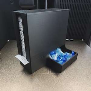 Può includere: Un dispenser rettangolare nero opaco dotato di una fessura verticale riempita con una pila di carte rettangolari bianche e un vassoio aperto collegato contenente una collezione di piccoli tappi in plastica blu traslucida.