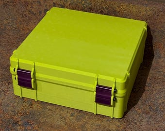 Puede incluir: Un estuche de almacenamiento cuadrado de color verde lima con una parte superior plana y un acabado mate liso. El contenedor cuenta con dos cierres rectangulares de color morado oscuro en la parte frontal, asegurados con pequeños pasadores negros.