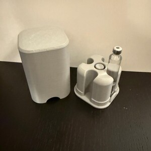 May include: A light gray, 3D-printed medical device set. The set includes a rectangular container with a lid and a holder with three circular openings, one holding a small glass vial. The objects are on a dark surface.
