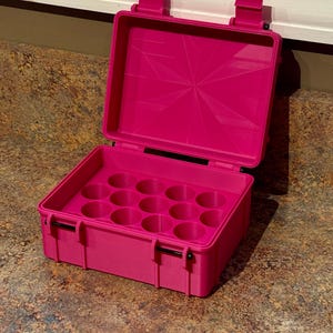 May include: A bright pink rectangular storage case with a hinged lid and two front latches. The interior features a molded insert with twelve circular compartments arranged in a grid, designed for holding small cylindrical items securely in place.