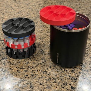 May include: A black insulated tumbler with a red lid next to a black and red 3D-printed organizer holding vials. The tumbler has the word "HYDRAPEAK" printed on it. The organizer is cylindrical with a grid-like top.