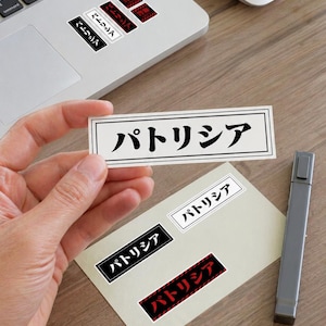 以下が含まれることがあります： 日本語で「パトリシア」と書かれたステッカーのコレクション。ステッカーは黒、白、赤など様々な色があります。一部のステッカーはノートパソコンに、その他は紙に貼られています。